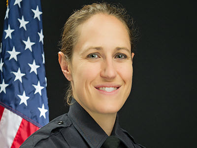 headshot of a smiling person in a black police uniform with a black background and an American flag in the background of the left side of the image
